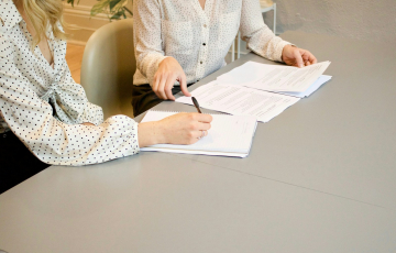 woman signing on white printer paper beside woman about to touch the documents