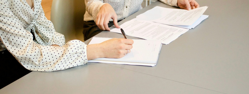 woman signing on white printer paper beside woman about to touch the documents